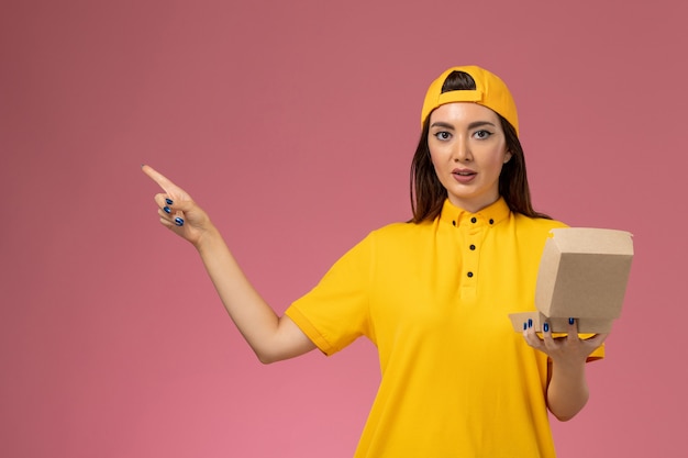 Free photo front view female courier in yellow uniform and cape holding little delivery food package on light-pink wall service uniform delivery company worker