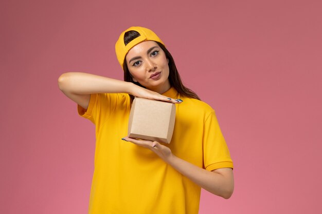 Front view female courier in yellow uniform and cape holding little delivery food package on light-pink wall uniform service delivery company girl