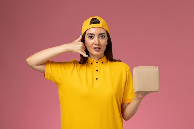 Front view female courier in yellow uniform and cape holding little delivery food package on light-pink wall uniform service delivery company job work