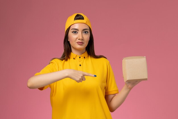 Front view female courier in yellow uniform and cape holding little delivery food package on light-pink wall uniform service delivery company