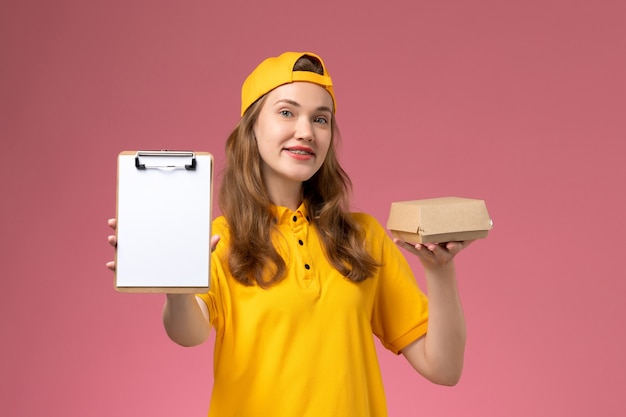 Free photo front view female courier in yellow uniform and cape holding little delivery food package and notepad on light pink wall service delivery uniform job