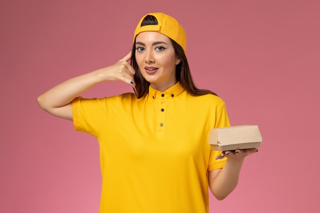 Front view female courier in yellow uniform and cape holding little delivery food package on the pink wall uniform service delivery company job