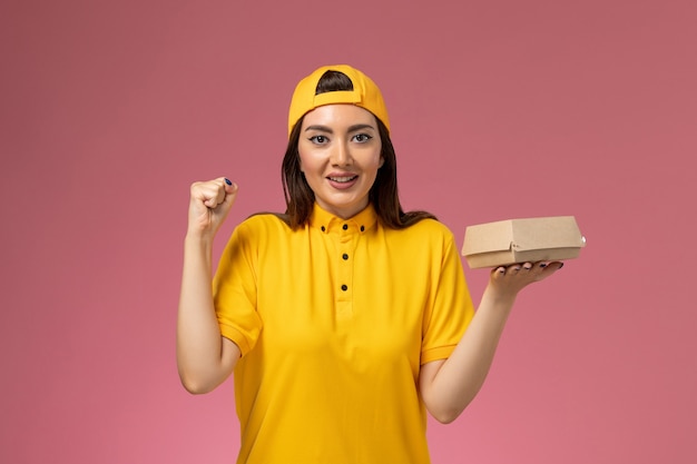 Front view female courier in yellow uniform and cape holding little delivery food package on the pink wall uniform service delivery company work