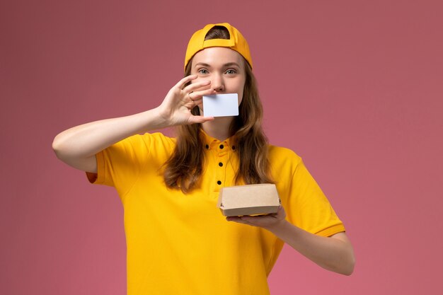 Front view female courier in yellow uniform and cape holding little delivery food package and plastic card on pink wall job service delivery worker