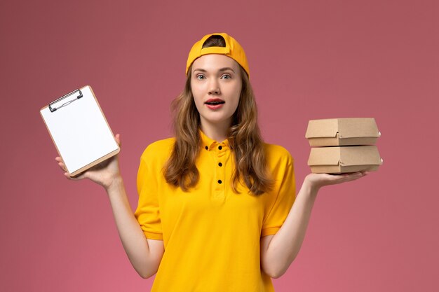 Front view female courier in yellow uniform and cape holding little delivery food packages with notepad on pink wall service delivery uniform job work