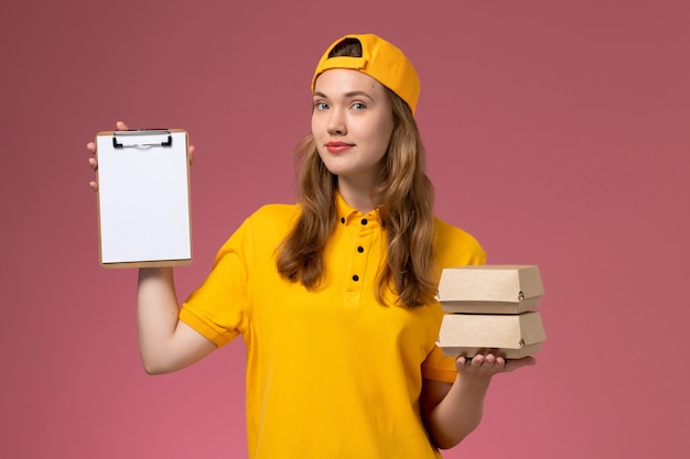 Front view female courier in yellow uniform and cape holding little delivery food packages with notepad on pink wall service delivery uniform