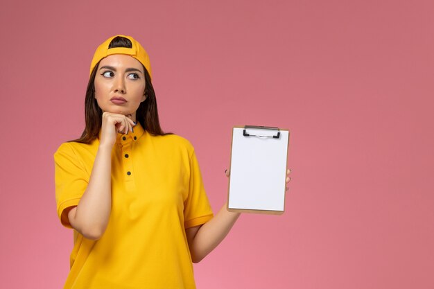 Front view female courier in yellow uniform and cape holding notepad thinking on pink wall company service uniform delivery