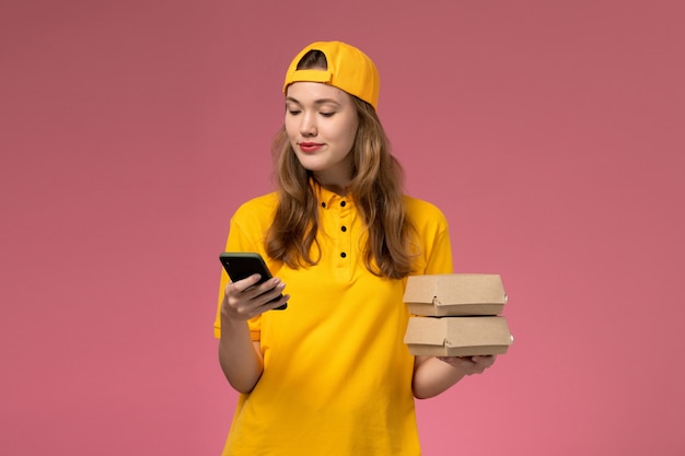 Front view female courier in yellow uniform and cape holding phone with food packages on pink wall company service delivery uniform work