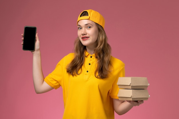 Front view female courier in yellow uniform and cape holding phone with food packages on pink wall company service delivery uniform