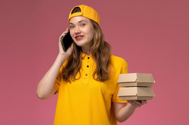 Front view female courier in yellow uniform and cape holding phone with food packages on pink wall company service delivery work uniform