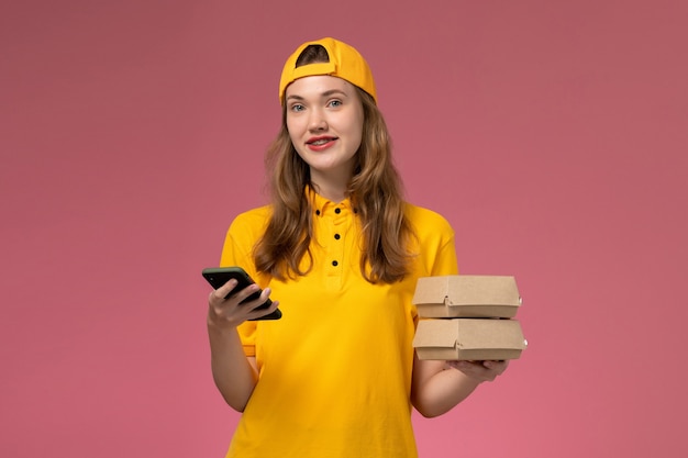 Front view female courier in yellow uniform and cape holding phone with food packages smiling on pink wall company service delivery uniform