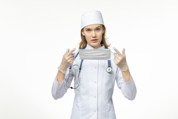 Front view female doctor in medical suit holding protective mask due to coronavirus on the white wall pandemic covid- virus illness