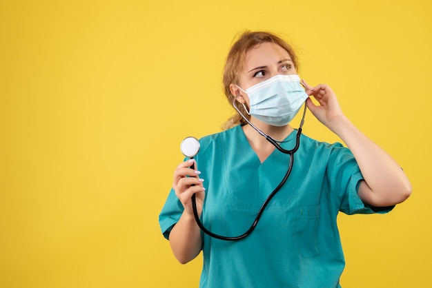 Front view of female doctor in medical suit and mask with stethoscope on yellow wall