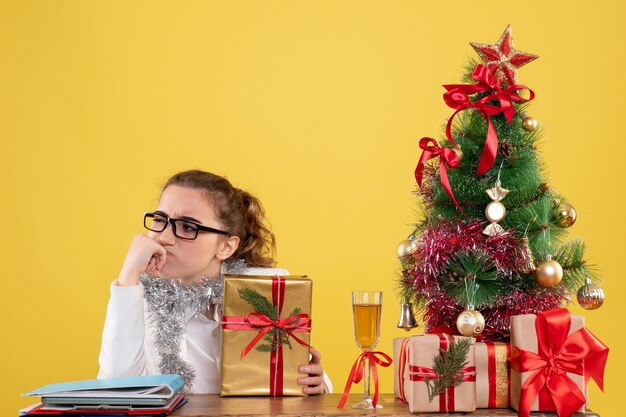 Front view female doctor sitting around christmas presents and tree thinking on light yellow background