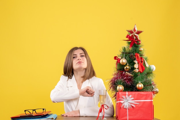 Free photo front view female doctor sitting in front of her table on yellow background with christmas tree and gift boxes