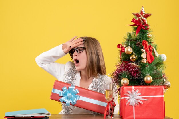 Front view female doctor sitting in front of table with presents and tree on the yellow background with christmas tree and gift boxes