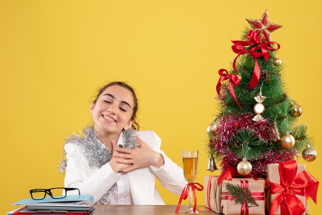 Front view female doctor sitting behind her table delighted on yellow background with christmas tree and gift boxes