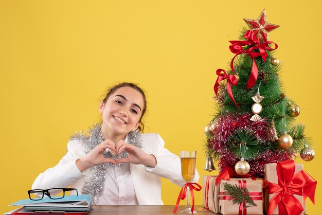 Front view female doctor sitting behind her table sending love on yellow background with christmas tree and gift boxes