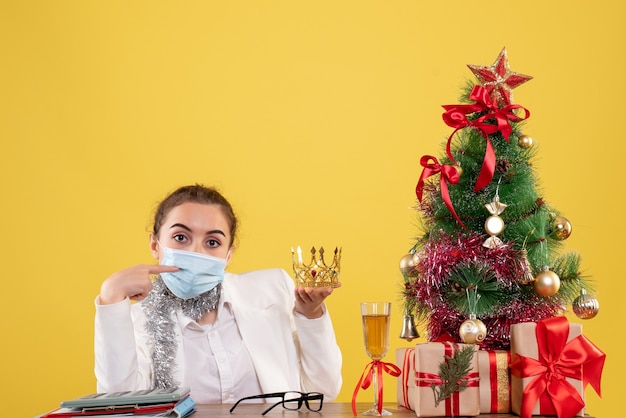 Front view female doctor sitting in sterile mask holding crown on yellow background with christmas tree and gift boxes