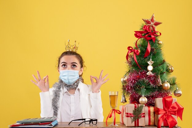 Front view female doctor sitting in sterile mask on yellow background with christmas tree and gift boxes