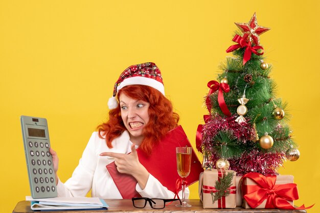 Free photo front view female doctor sitting behind table and holding calculator on yellow background with christmas tree and gift boxes