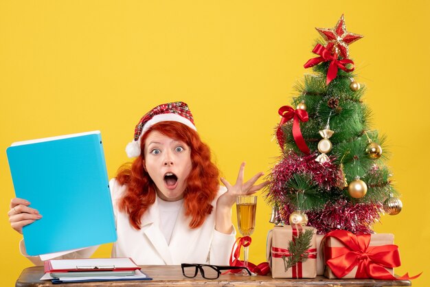 Front view female doctor sitting behind table and holding documents on a yellow background with christmas tree and gift boxes