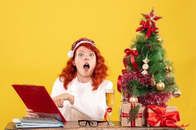 Front view female doctor sitting behind table shockingly reading notes on yellow background with christmas tree and gift boxes