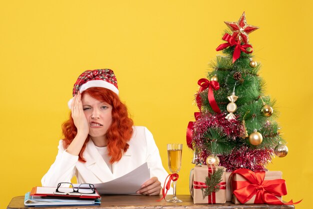 Front view female doctor sitting behind table with presents and documents on yellow background  with christmas tree and gift boxes