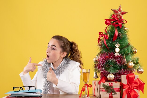 Front view female doctor sitting behind table with xmas presents and tree on yellow background