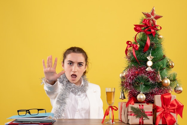 Front view female doctor sitting behind table on yellow background with christmas tree and gift boxes