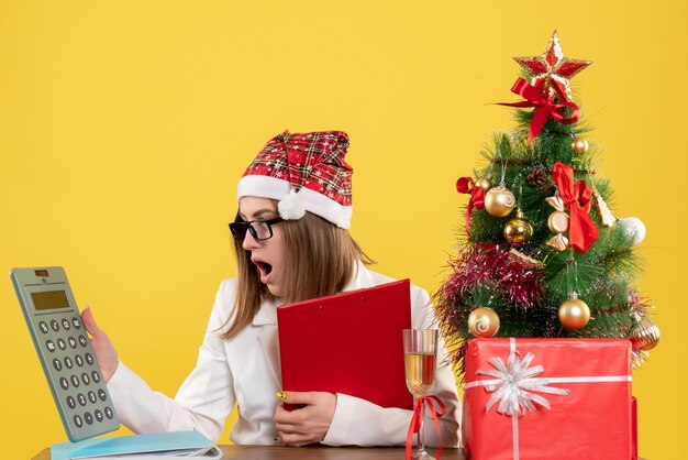 Front view female doctor sitting with xmas presents holding calculator on yellow background