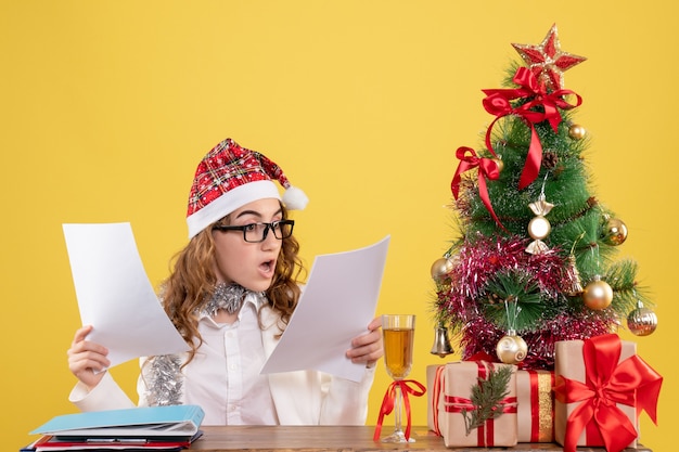 Front view female doctor sitting with xmas presents tree and holding documents on yellow background