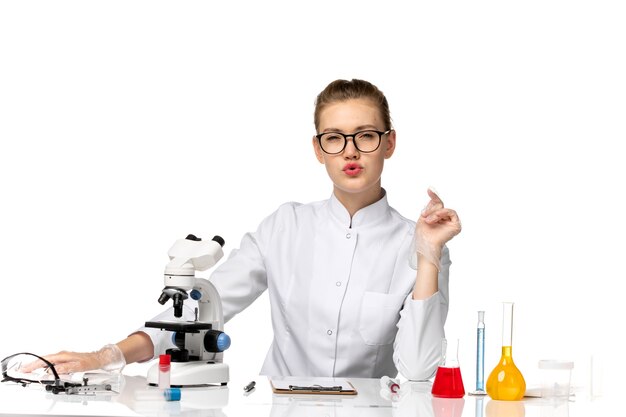 Front view female doctor in white medical suit sitting in front of table with solutions on light white space