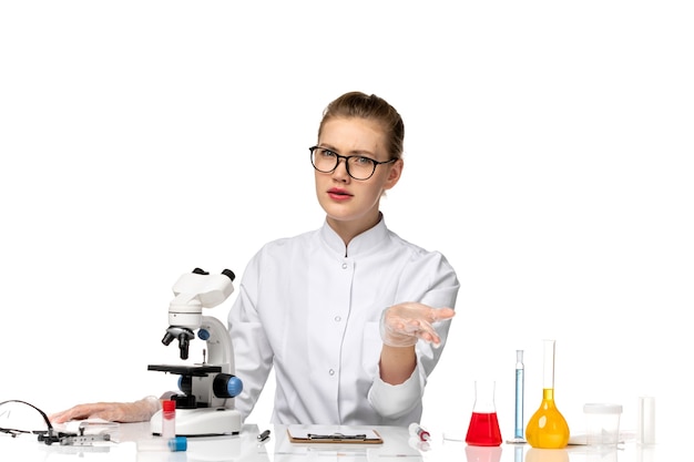 Front view female doctor in white medical suit sitting in front of table with solutions on a white space