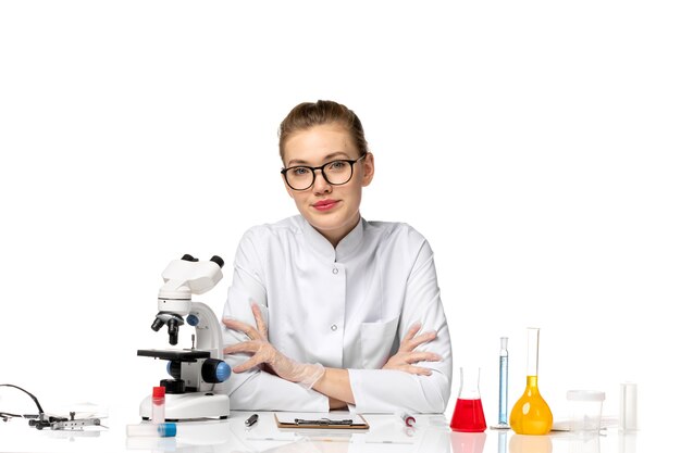 Front view female doctor in white medical suit sitting in front of table with solutions on a white space