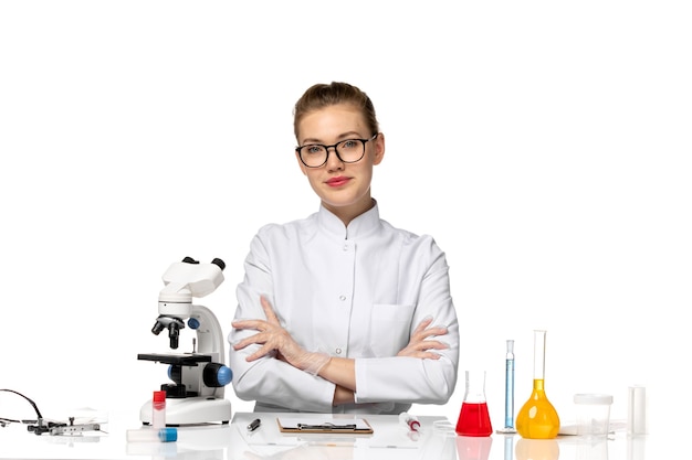 Front view female doctor in white medical suit sitting in front of table with solutions on white space