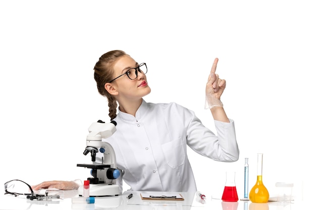 Front view female doctor in white medical suit sitting in front of table with solutions on white space