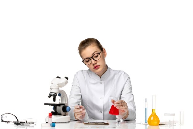 Front view female doctor in white medical suit with gloves holding flask with solution on light white space