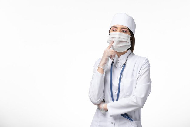 Front view female doctor in white medical suit with a mask due to coronavirus deeply thinking on white wall health disease pandemic covid-