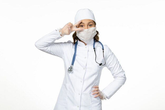 Front view female doctor in white medical suit and with mask due to coronavirus on the white wall virus disease pandemic covid