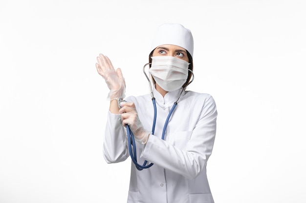 Front view female doctor in white medical suit with a mask due to pandemic using stethoscope on white wall disease health medicine pandemic covid-