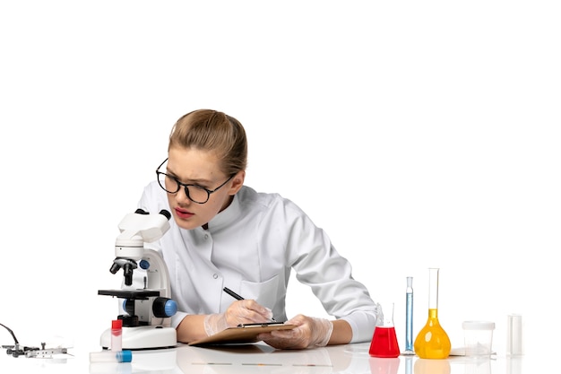 Front view female doctor in white medical suit working with microscope on a white space