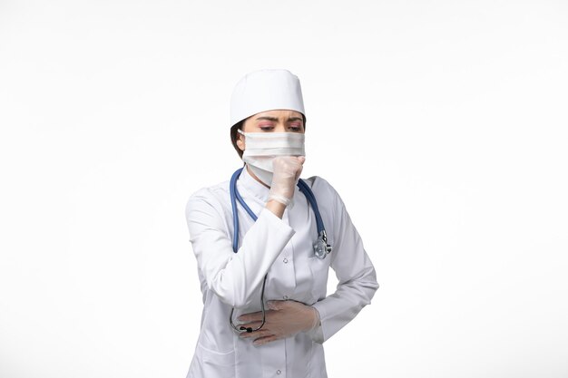 Front view female doctor in white sterile medical suit and wearing mask due to covid- coughing on white wall illness virus covid- pandemic disease