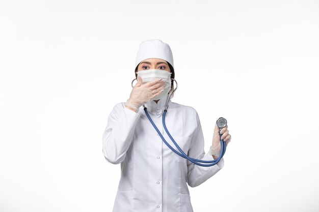 Front view female doctor in white sterile medical suit and wearing mask due to covid- using a stethoscope on light white wall illness virus covid- pandemic disease