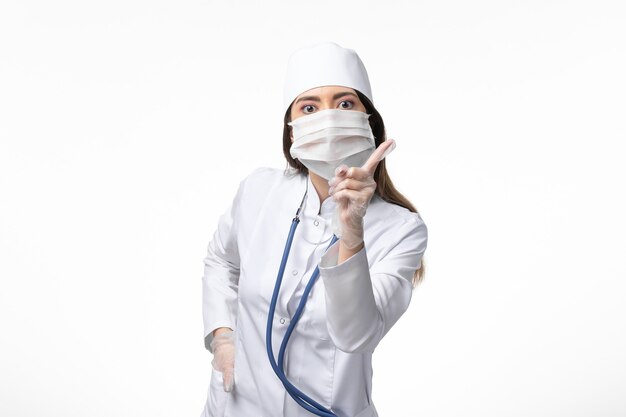 Front view female doctor in white sterile medical suit with mask due to coronavirus on the white wall illness covid- pandemic disease virus