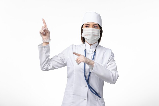 Front view female doctor in white sterile medical suit with mask due to coronavirus on white wall illness pandemic disease covid-