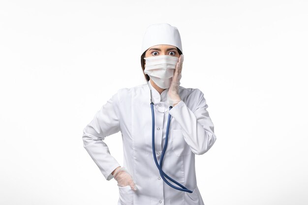 Front view female doctor in white sterile medical suit with mask due to coronavirus with shocked expression on white wall disease illness pandemic virus covid-