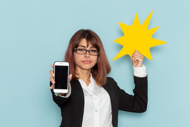 Front view female office worker in strict suit holding phone and yellow sign on blue surface