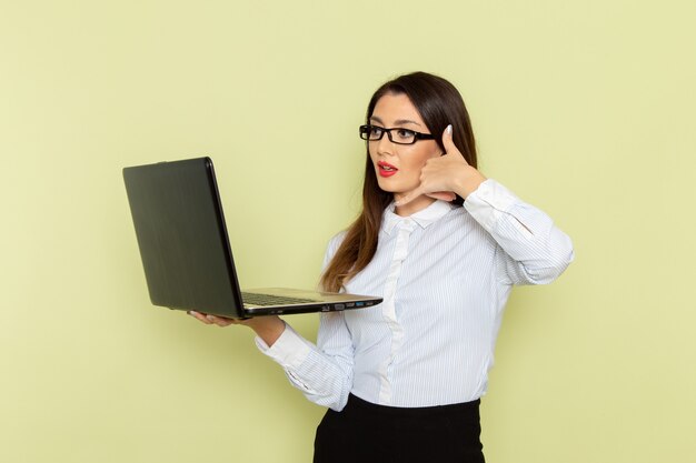 Front view of female office worker in white shirt and black skirt using her laptop on light-green wall