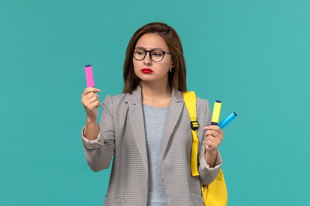 Front view of female student in grey jacket wearing her yellow backpack holding felt pens on light blue wall